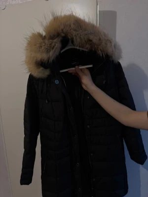 Svart lång dunjacka med fuskpäls - Snygg svart lång dunjacka från Boomerang med stor huva och fuskpälsdetalj. Jackan har dragkedja och knappar framtill, två sidofickor och quiltad design. Perfekt för kalla vinterdagar och ger en stilren look.