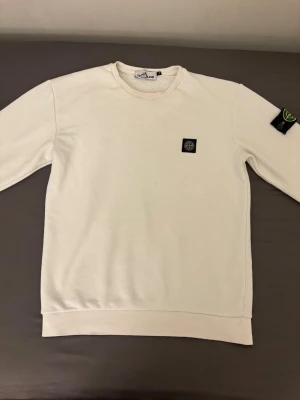 Vit sweatshirt från Stone Island - Vit sweatshirt från Stone Island med klassisk rund halsringning och ikonisk logopatch på vänster ärm. Tröjan har en rak passform och är tillverkad i mjuk bomull för maximal komfort. Minimalistisk design med diskret logga på bröstet.