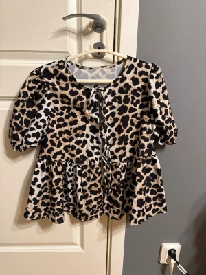 Leopardmönstrad blus med knyt - Snygg blus med leopardmönster och puffiga halvlånga ärmar. Blusen har volang nertill och knytning framtill vid halsen. Aldrig använt