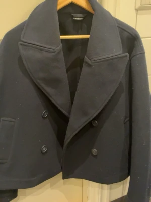 Marinblå skepparkavaj Gina Tricot XS - Stilren marinblå skepparkavaj från Gina Tricot i storlek XS. Kavajen har dubbelknäppning, breda slag och två fickor framtill. Klassisk kort modell i ullblandning som passar perfekt till kalla dagar.