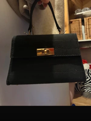 Svart handväska med krokoprägling - Stilren svart handväska med krokodilpräglat mönster och guldfärgat vridlås. Väskan har ett klassiskt handtag och en elegant, fyrkantig form. Perfekt accessoar för att lyfta din outfit.