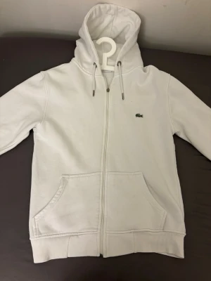 Vit zip hoodie från Lacoste - Snygg vit hoodie från Lacoste med dragkedja framtill och klassisk krokodil-logga på bröstet. Hoodien har huva med dragsko, ribbade muddar och en stor magficka. Perfekt för en clean och stilren look. Materialet känns mjukt och bekvämt.