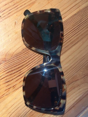 Bruna leopardmönstrade solglasögon - Snygga solglasögon från Ralph Lauren med leopardmönstrad båge i bruna och beige toner. Stora, fyrkantiga glas och mörka linser ger en cool look. Märkets logga i silver på skalmen. Perfekta för att lyfta din stil i solen.