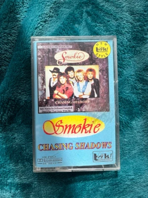 Rare old collectible cassette-Smokie Chasing Shadows kassettband - Säljer ett klassiskt kassettband med albumet Chasing Shadows av Smokie. Omslaget är blått med bandets logga och bild på medlemmarna. Kassetten är svart och genomskinlig med etikett från Laki Records. Perfekt för dig som samlar på retro musikprylar.