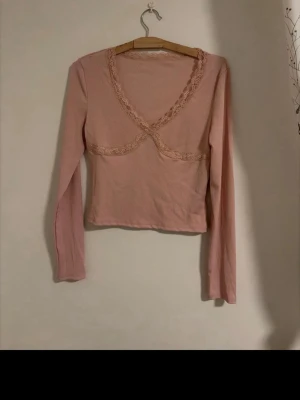 Rosa långärmad topp med spetsdetaljer - Söt ljusrosa långärmad topp med v-ringning och fina spetsdetaljer längs halsringningen och under bysten. Oanvänd utan prislapp