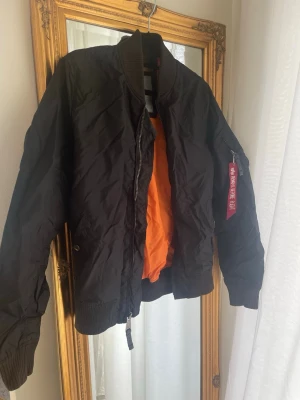 Svart bomberjacka från Alpha Industries - Klassisk svart bomberjacka från Alpha Industries med orange foder och ribbade muddar. Jackan har dragkedja, fickor med tryckknappar och en ikonisk röd tag med texten 'Remove Before Flight' på ärmen. Perfekt för en streetwear-look. Storlek L kan även passa M beroende på hur man vill att den ska sitta 