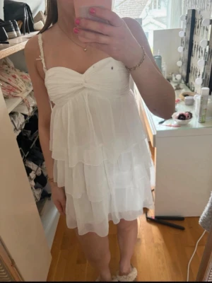 Vit volangklänning med smala axelband - Söt vit kortklänning med flera lager volanger och smala axelband. Klänningen har en twistad detalj vid bysten och är tillverkad i ett lätt, luftigt material som ger en romantisk känsla.