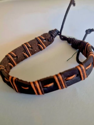 Brunt läderarmband med orange snören - Snyggt armband i brunt läder med orange snören lindade runt bandet. Justerbar storlek med knytning för perfekt passform. Perfekt accessoar för en avslappnad och trendig stil.