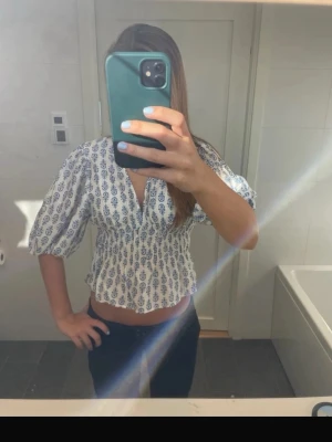 Vit och blå blus från H&M, S - Supersöt blus från H&M i vitt med blått mönster. Blusen har v-ringning, smockad midja och puffiga trekvartsärmar. Perfekt till jeans eller kjol för en fräsch look. Materialet är mjukt och lätt, troligtvis bomull eller polyester.