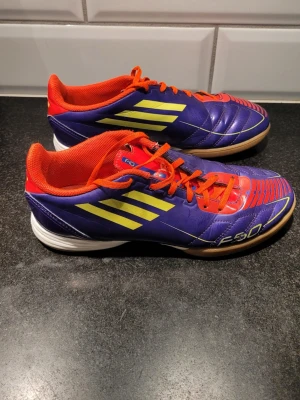 Adidas F50 lila inomhusfotbollsskor - Säljer ett par Adidas F50 inomhusfotbollsskor i lila med gula ränder och röda detaljer. Skorna har en låg profil, snörning och slitstark gummisula för bra grepp på inomhusplan. Ovandelen är i syntetmaterial och insidan är fodrad i rött tyg. Perfekta för snabba vändningar på futsal eller inomhusfotboll. Väldigt fint skick!