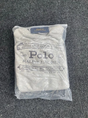 Grå hoodie från Polo Ralph Lauren - Snygg grå hoodie från Polo Ralph Lauren med tryck på bröstet och klassisk logga. Hoodien har huva med dragsko, ribbade muddar och en stor magficka. Perfekt för dig som gillar stilren streetwear och vill ha något bekvämt och trendigt.