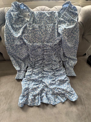 Blå blommig kortklänning med puffärm - Supersöt blå kortklänning med vita blommor och volang längst ner. Klänningen har puffiga långärmar med smock vid ärmslut och rynkade detaljer längs hela kroppen. Perfekt för dig som gillar romantiska och trendiga plagg.