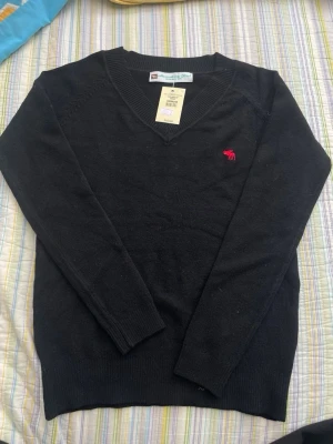Svart cashmere tröja Abercrombie & Fitch - Svart stickad tröja från Abercrombie & Fitch med V-ringning och röd broderad älg-logga på bröstet. Tröjan har långa ärmar och ribbade muddar vid hals, ärmslut och nederkant. Perfekt för en stilren och enkel look. 100% cashmere