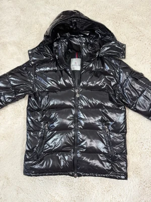 Svart glansig dunjacka från Moncler - Snygg svart dunjacka från Moncler med glansig finish och huva. Jackan har två dragkedjefickor framtill, quiltad design och hög krage. Perfekt för kalla vinterdagar och ger en cool streetstyle-look.