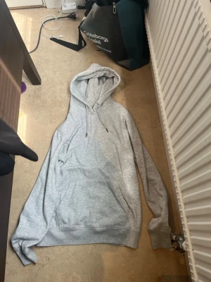 Ljusgrå hoodie med huva - Säljer en stilren ljusgrå hoodie med huva och dragsnören. Hoodien har en stor magficka och ribbade muddar vid ärmslut och nederkant. Perfekt för en avslappnad och trendig look. S men passar mer M och L