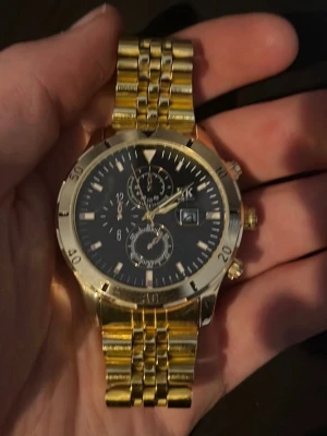 Guldfärgad kronografklocka med svart urtavla - Stilren klocka med guldigt metallarmband och svart urtavla. Klockan har kronograf-funktioner, datumvisning och tydliga markeringar. Perfekt accessoar för dig som vill sticka ut med en lyxig look.