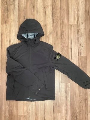Svart vindjacka från Stone Island - Svart vindjacka från Stone Island med huva och dragkedja framtill. Jackan har en ikonisk logotyp-patch på vänster ärm och är tillverkad i nylon och Tyvek-material. Perfekt för blåsiga dagar och har en modern, sportig look.