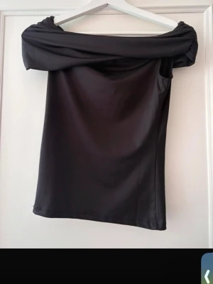 Svart offshoulder topp - Snygg svart offshoulder topp med bred krage som vilar över axlarna. Toppens material har en lätt glans och känns mjukt mot huden. Perfekt för dig som vill ha en stilren och trendig look.