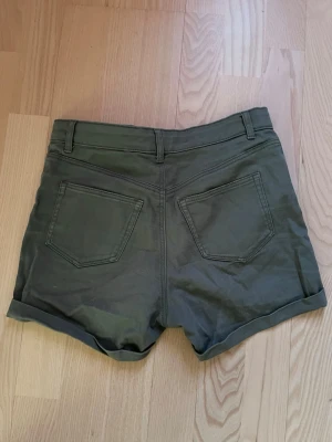 Olivgröna high waist shorts - Snygga olivgröna shorts med hög midja och uppvikta benslut. Klassisk femficksmodell med dragkedja och knapp framtill samt bälteshällor. Perfekta för varma dagar och enkel att matcha med olika toppar.