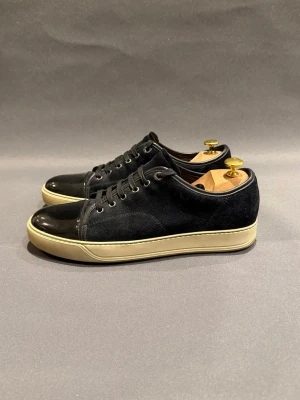 Svarta Lanvin sneakers med mocka och lack - Stilrena svarta sneakers med ovandel i mocka och lackdetaljer på tån. Skorna har vita sulor och klassisk snörning. Perfekta för dig som gillar en clean och modern look med lite extra edge.