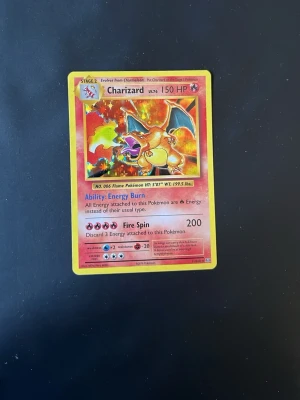 Charizard Pokémonkort Holo - Säljer ett Charizard Pokémonkort med holografisk framsida. Kortet har en gul ram, färgglad illustration och text på engelska. Baksidan har klassiskt blått Pokémon-motiv med pokéboll. Perfekt för samlare eller Pokémonfans.