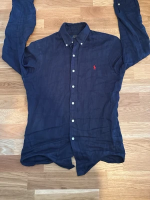 Mörkblå linneskjorta Polo Ralph Lauren - Snygg mörkblå skjorta i linne från Polo Ralph Lauren med röd broderad logga på bröstet. Slim fit-modell med klassisk krage, vita knappar och långa ärmar. Perfekt för en stilren och avslappnad look. Storleken är i slimfit XS och skjortan har inga defekter! 