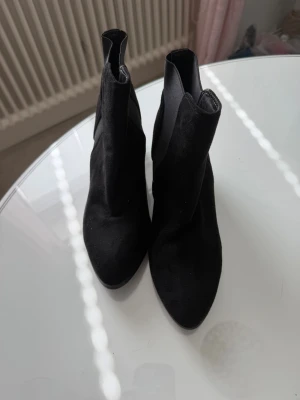 Svarta stövletter med klack - Säljer ett par svarta stövletter med rundad tå och bred klack. Skorna är i mockaimitation och har en stilren, minimalistisk design utan synliga detaljer. Perfekta för dig som gillar en clean look och vill ha något som funkar till många outfits.