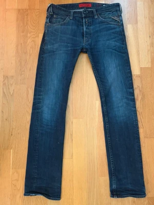 Mörkblå vintage jeans från Replay - Snygga mörkblå jeans från Replay med klassisk femficksmodell och diskreta slitningar. Jeansen har raka ben och en röd detalj på linningen. Perfekta för en trendig och avslappnad stil.