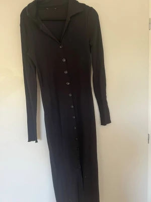 Svart långärmad skjortklänning - Snygg svart långklänning med långa ärmar och knappar längs hela framsidan. Klänningen har en krage och är figurnära i modellen, perfekt för dig som gillar en stilren look.