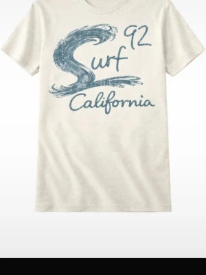 Vit t-shirt med blått surftryck - Snygg vit t-shirt med blått tryck som föreställer en våg och texten 'Surf 92 California' på framsidan. Klassisk passform och rund halsringning, perfekt för en avslappnad stil.
