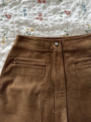 Brun äkta mocka med fickdetaljer - Snygg brun kjol i mocka med två fickdetaljer framtill och knapp i midjan. Kjolen har en klassisk A-linjeform och är perfekt för dig som gillar en stilren och trendig look. UK4 passar xxs och xs