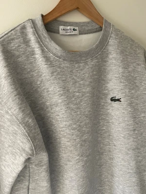 Grå sweatshirt från Lacoste - Klassisk grå sweatshirt från Lacoste med rund halsringning och broderad krokodil-logga på bröstet. Tröjan har en relaxed fit och är tillverkad i mjukt bomullsmaterial. Perfekt för en avslappnad och stilren look.