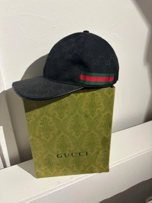 Svart keps från Gucci med logomönster - Svart keps från Gucci med diskret GG-logomönster över hela kepsen och klassisk böjd skärm. På sidan finns en ikonisk grön och röd rand. Kepsen är tillverkad i canvasmaterial och har en stilren, modern look.