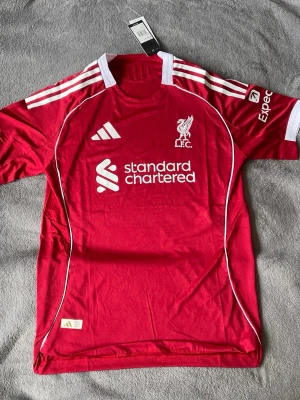 Liverpool 24/25 Home - Riktigt clean Liverpool-tröja i nya hemmadesignen 😮‍💨 Klassisk röd med vita detaljer — ser sjukt bra ut både till match och som outfit.  • Liverpool FC • 24/25 Home Kit • Storlek M (L men passar som M) • Standard Chartered sponsor • Ny med tags • Skön passform & fina detaljer • Perfekt för alla Liverpool-fans 🔴  En av de snyggaste Liverpool-tröjorna på länge enligt mig 👀  💰 Pris: 449 kr 📍  skickas  