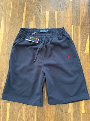 Marinblå shorts Polo Ralph Lauren - Snygga marinblå shorts från Polo Ralph Lauren med elastisk midja och dragsko. Klassisk röd logga broderad på vänster ben. Tillverkade i mjukt sweatshirtmaterial för en avslappnad look. Perfekta för chill dagar eller sportiga outfits.