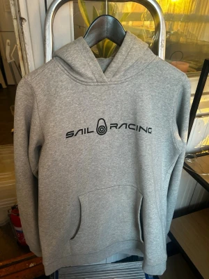Grå hoodie från Sail Racing - Säljer en stilren grå hoodie från Sail Racing med svart logga tryckt på bröstet. Hoodien har en klassisk huva och en stor magficka framtill. Tillverkad i mjukt sweatshirtmaterial som är perfekt för chill dagar. Passar dig som gillar enkel och sportig stil.