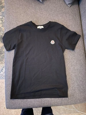 Svart Moncler t shirt  - Snygg svart t-shirt från Moncler med klassisk rund hals och korta ärmar. På bröstet sitter Monclers ikoniska logga som en patch. Perfekt basplagg i mjuk bomull som funkar till allt.