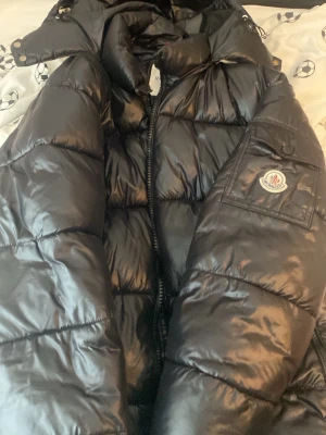Svart dunjacka från Moncler - Snygg svart dunjacka från Moncler med klassisk glansig finish och broderad logga på ärmen. Jackan har hög krage, dragkedja framtill och quiltad design som håller värmen. Perfekt för kalla vinterdagar och riktigt stilren look.