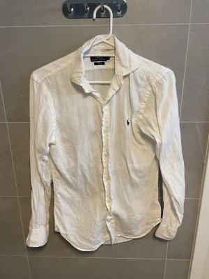 Vit linneskjorta från Ralph Lauren - Klassisk vit skjorta i linne från Ralph Lauren med lång ärm och knappar framtill. Skjortan har en broderad logga på bröstet och en avslappnad passform. Perfekt för en stilren och fräsch look. Jävligt dåliga bilder! Orkade inte ta bättre. skriv ifall bra bilder krävs. Lite skrinkliga o så men det försvinner i tvätten eller om du ska strika dom eller hur du än gör