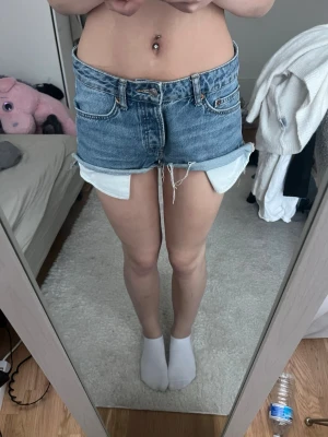 Blå denimshorts med uppvik - Snygga blå jeansshorts med råa kanter och uppvikta ben. Klassisk femficksmodell med dragkedja och knapp framtill. Perfekta för varma dagar och enkel att matcha med allt.