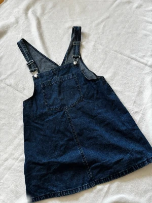 Mörkblå jeansdress med hängslen - Snygg mörkblå jeans klänning/dress i klassisk denim med justerbara hängslen och en stor bröstficka framtill. Rak passform och kort modell som passar perfekt till en avslappnad look.