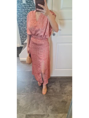 Rosa långklänning med zebramönster - Snygg rosa långklänning med zebramönster och v-ringning. Klänningen har lös passform upptill, markerad midja och en hög slits framtill. Perfekt för sommarens alla tillfällen.