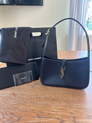 Ysl väska - Saint Laurent Le 5 À 7 shoulder bag in black smooth calfskin leather with gold-tone YSL hardware. Purchased from Farfetch and only used a few times. Retail price: 2205€ / 24000SEK  Condition is very good with only a few very small/light marks from normal wear (see pictures) The leather and structure are still beautiful, and the bag has been well cared for.  Comes with: 	•	Original Saint Laurent dust bag 	•	Saint Laurent shopping bag 	•	Authenticity/cards received with     purchase 	•	Original pa