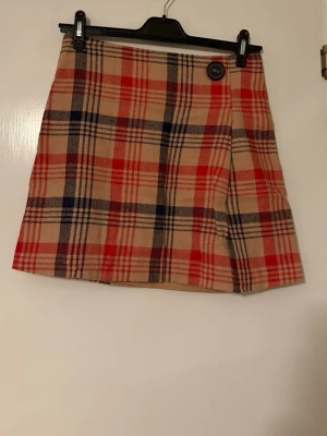 Rutig kortkjol från Holly & Whyte Lindex - Snygg rutig kortkjol från Holly & Whyte by Lindex i beige, rött och svart. Kjolen har en klassisk A-linjeform och ett dekorativt knappdetalj framtill. Perfekt för dig som gillar preppy stil och vill sticka ut lite extra.