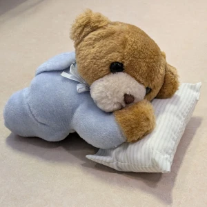 Söt björn liggande på en kudde - Jätte söt och mjuk leksaksbjörn i blå pyjamas som ligger på en kudde. Produkten är i gott skick utan synligt slitage. Längd ca 20 cm