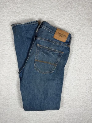 Abercrombie & Fitch jeans storlek W31 L32 - Snygga blå jeans från Abercrombie & Fitch med klassisk femficksdesign och raka ben. Jeansen har en läderpatch med logga bak i midjan och subtila slitningar framtill. Perfekta för en avslappnad och trendig look.