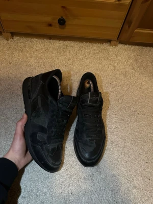 Valentino rockrunners  - Säljer ett par svarta sneakers från Valentino med mörkgrönt camomönster. Skorna har låga skaft, klassisk snörning och är tillverkade i en mix av mocka och textil. Men tyvärr lite hål i hälen efter andvändning. Snygg och stilren design med diskret logga på plösen. Perfekta för dig som gillar exklusiva skor. Pris går att diskutera.