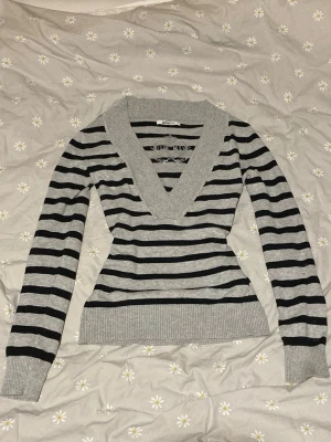 Gråmelerad randig tröja Gina Tricot - Stickad tröja från Gina Tricot i gråmelerat med svarta horisontella ränder. Djup v-ringning med bred ribbad kant och broderad logga på bröstet. Långa ärmar och ribbade muddar. Perfekt för en avslappnad men snygg look.