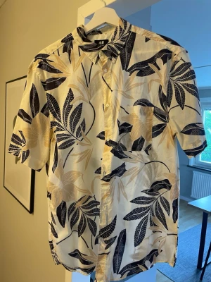 Blommig kortärmad skjorta från H&M strlk L - Snygg kortärmad skjorta från H&M med blommigt mönster i mörkblått och beige på vit botten. Skjortan har klassisk krage, knappar framtill och en bröstficka. Perfekt för sommarens alla tillfällen!