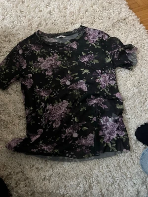 Svart meshblus med lila blommor - Snygg svart meshblus med lila blommigt mönster. Blusen är genomskinlig och rund halsringning. Perfekt att styla lager på lager för en trendig look.